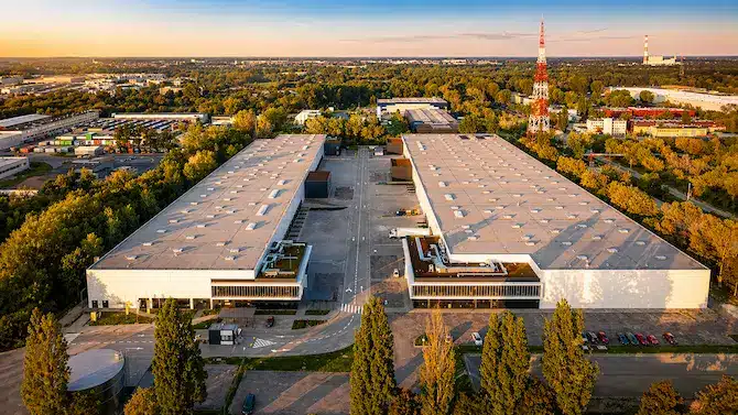 Aerial view of a large industrial complex with two long parallel warehouse buildings separated by a central road, surrounded by trees and parking lots.